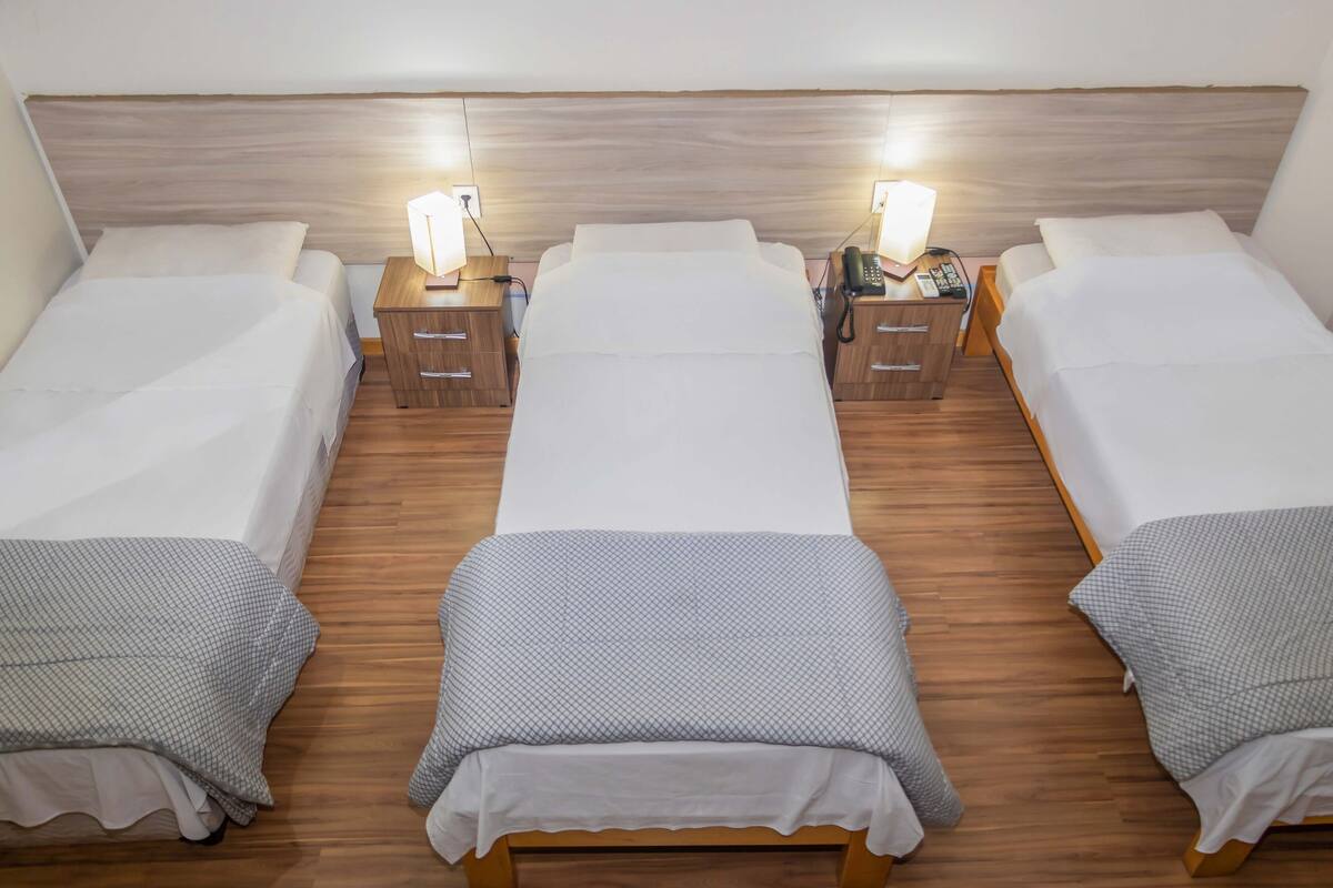 A hotel room with three single beds, wooden headboards, bedside tables with lamps, and a wooden floor.