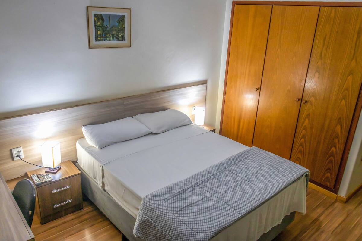 A neatly made bed with white linens, a bedside table with a lamp, a small drawer, and a wooden wardrobe.