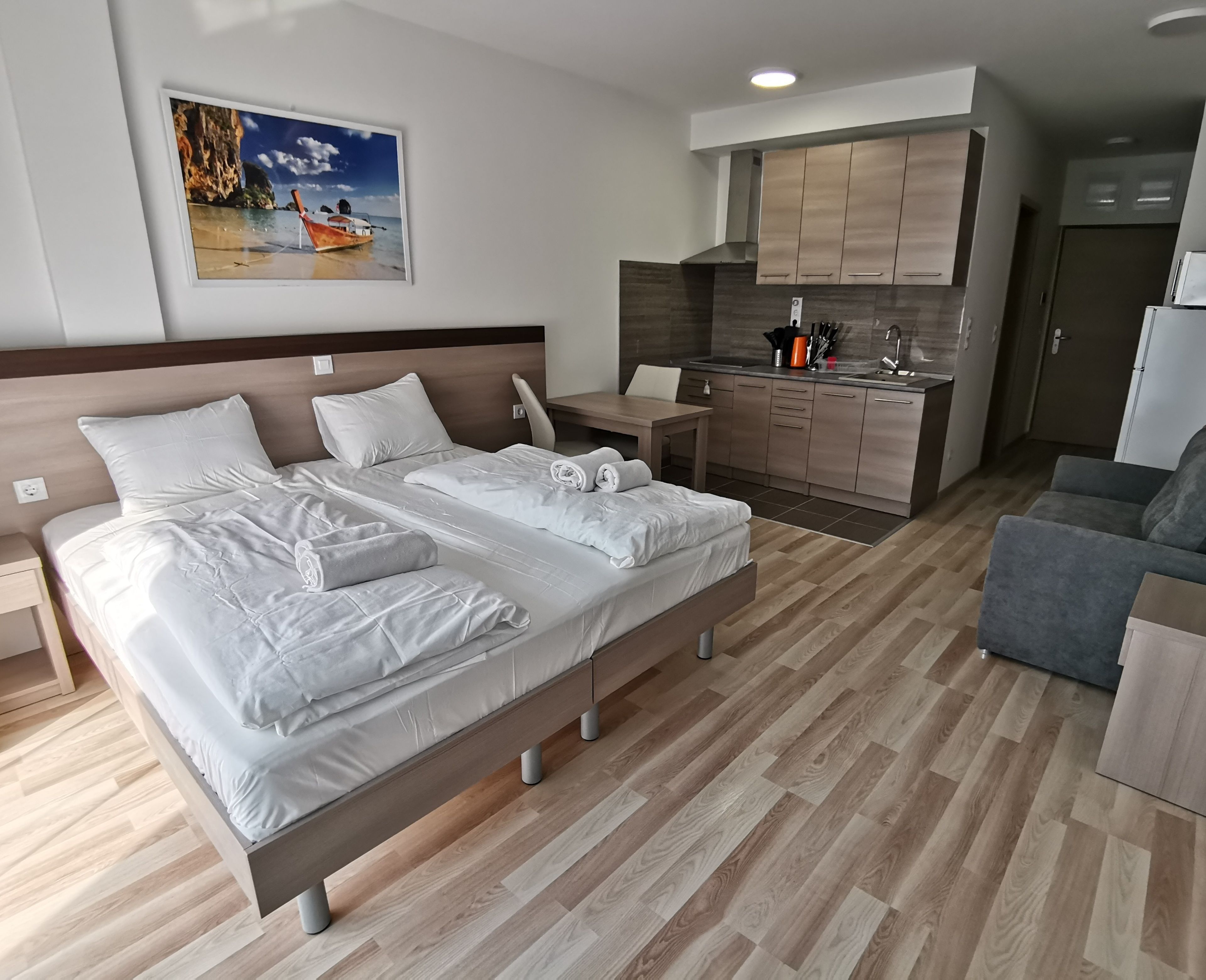 A modern hotel room with a large bed, a small desk, a kitchenette, and a view of a boat on the beach.
