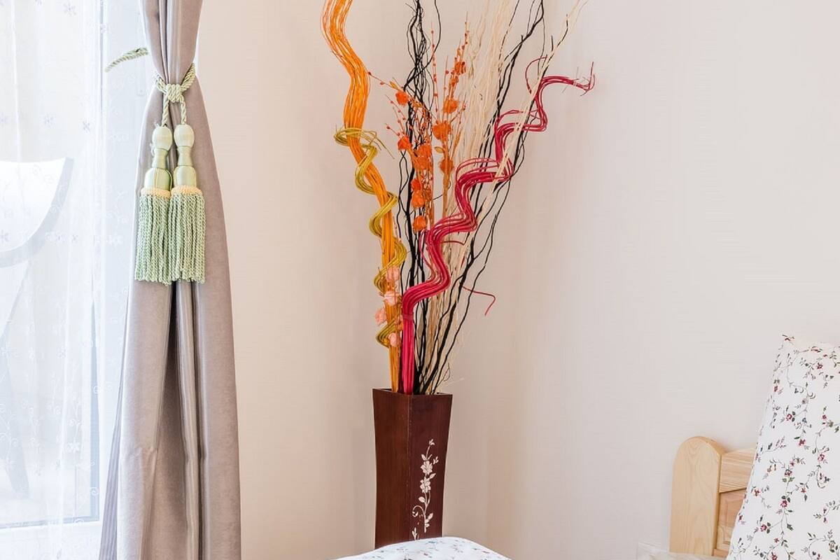 Een net opgemaakt bed met een bloemenprintdeken, een vaas met decoratieve takken en een kwast aan het gordijn.