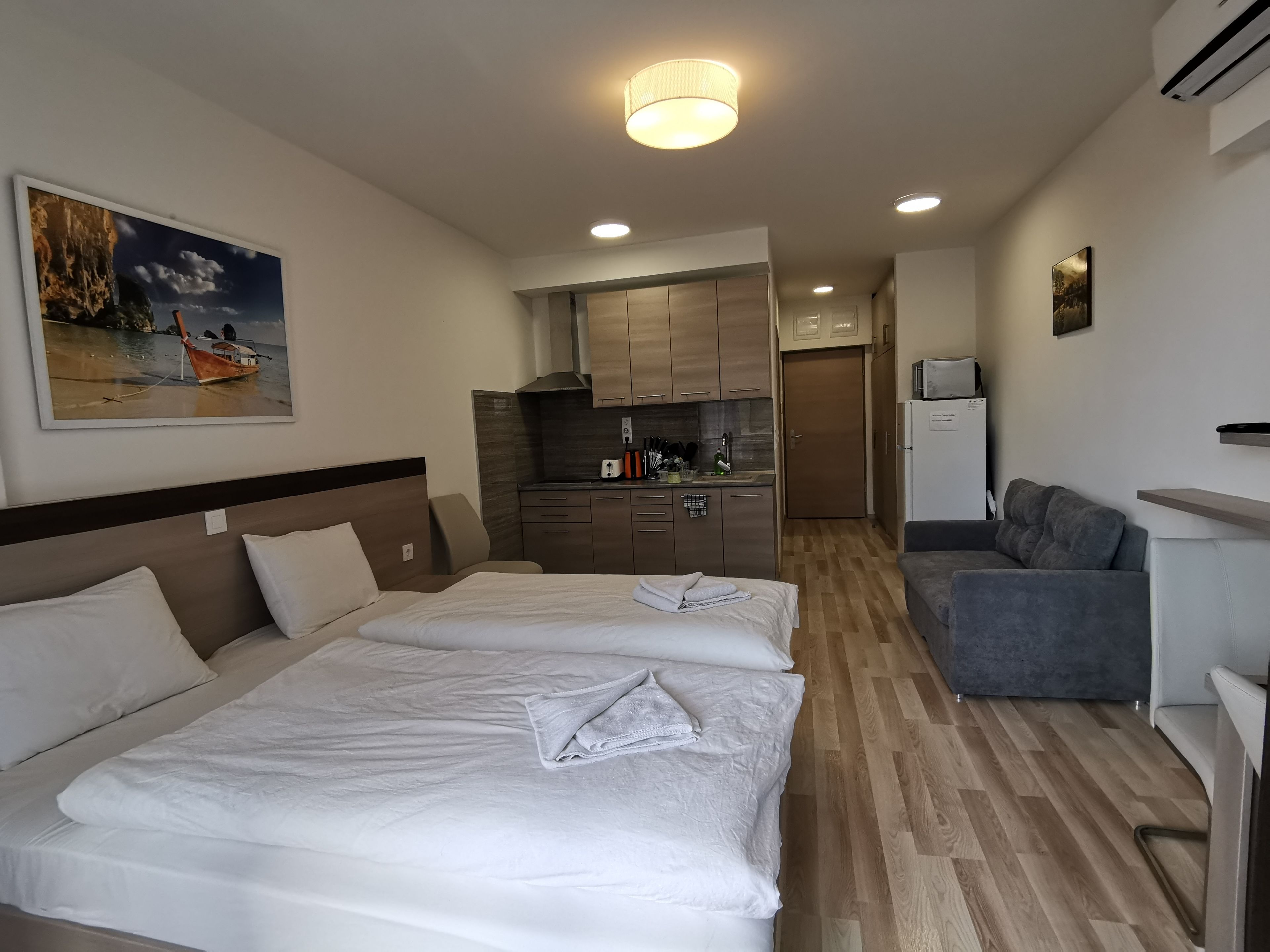 A hotel room with two beds, a kitchenette, a gray sofa, and a painting on the wall.