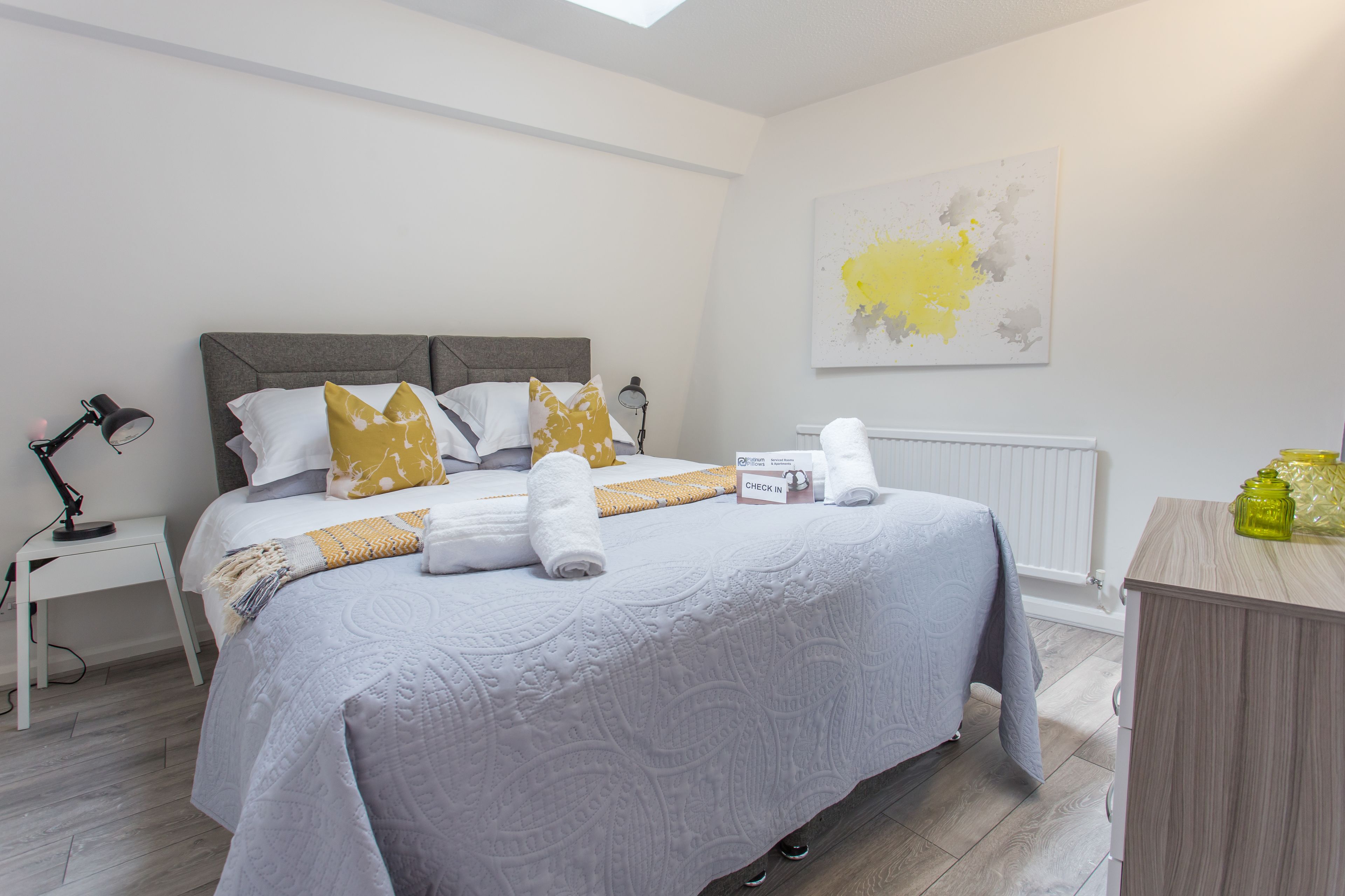 Un letto rifatto con cura, con testiera grigia, biancheria bianca e coperta gialla. Si nota un comodino con una lampada, un termosifone bianco e un quadro astratto appeso al muro.