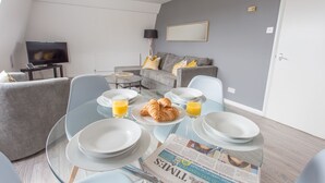 A modern living room with a dining area, grey sofas, a flat-screen TV, and breakfast items on the table. - The Manor Suite (Oxford)