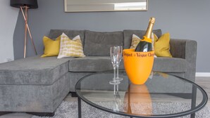 A modern living room with a grey sectional sofa, a glass coffee table, a bottle of champagne, and a glass. - The Manor Suite (Oxford)