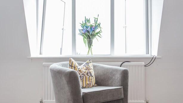 A grey armchair with a patterned pillow in front of a window with a white radiator. - The Manor Suite (Oxford)