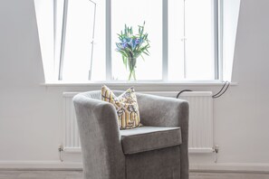 A grey armchair with a patterned pillow in front of a window with a white radiator. - The Manor Suite (Oxford)