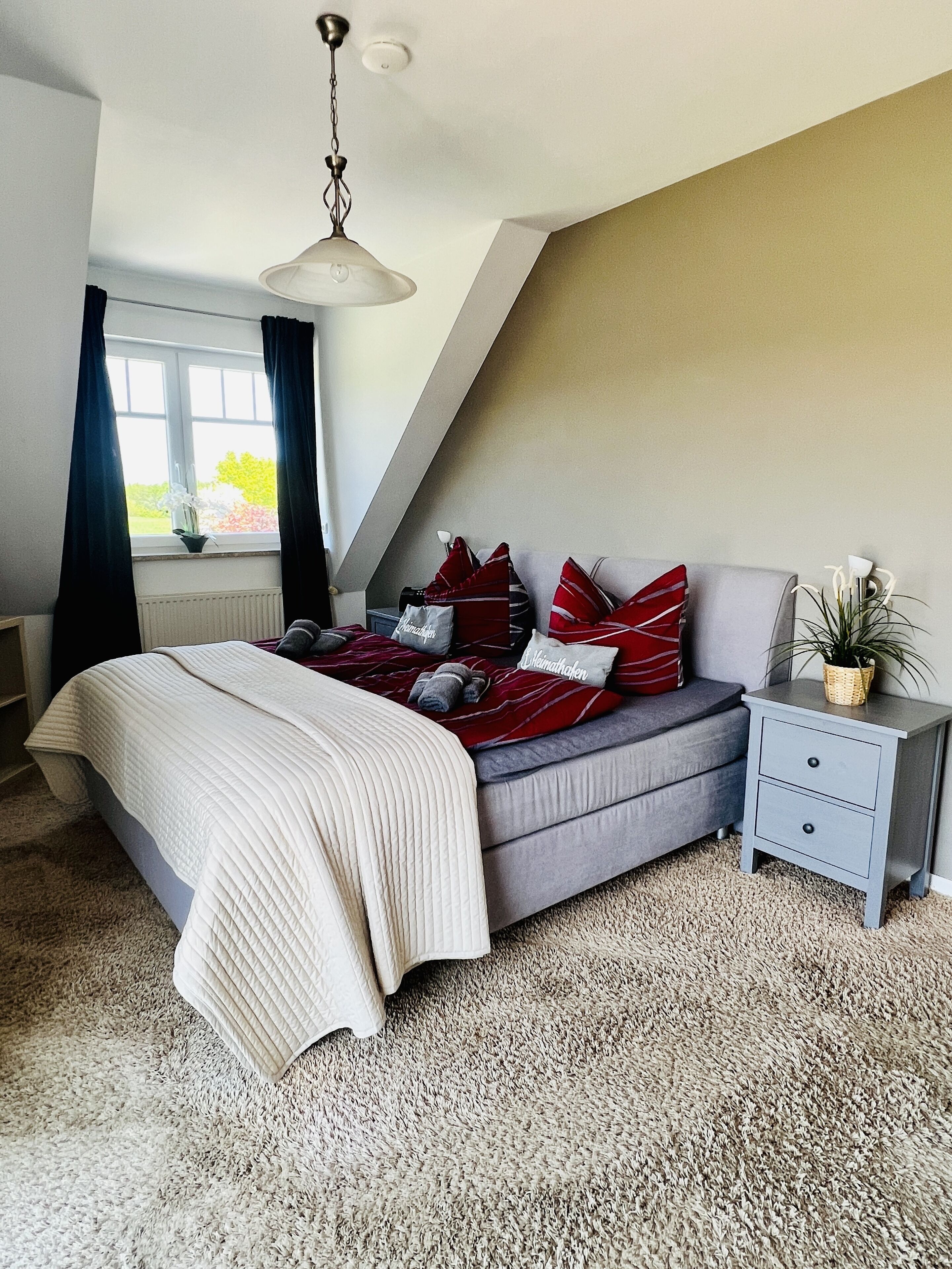 A bedroom with a bed, a nightstand, a window with curtains, and a plant.