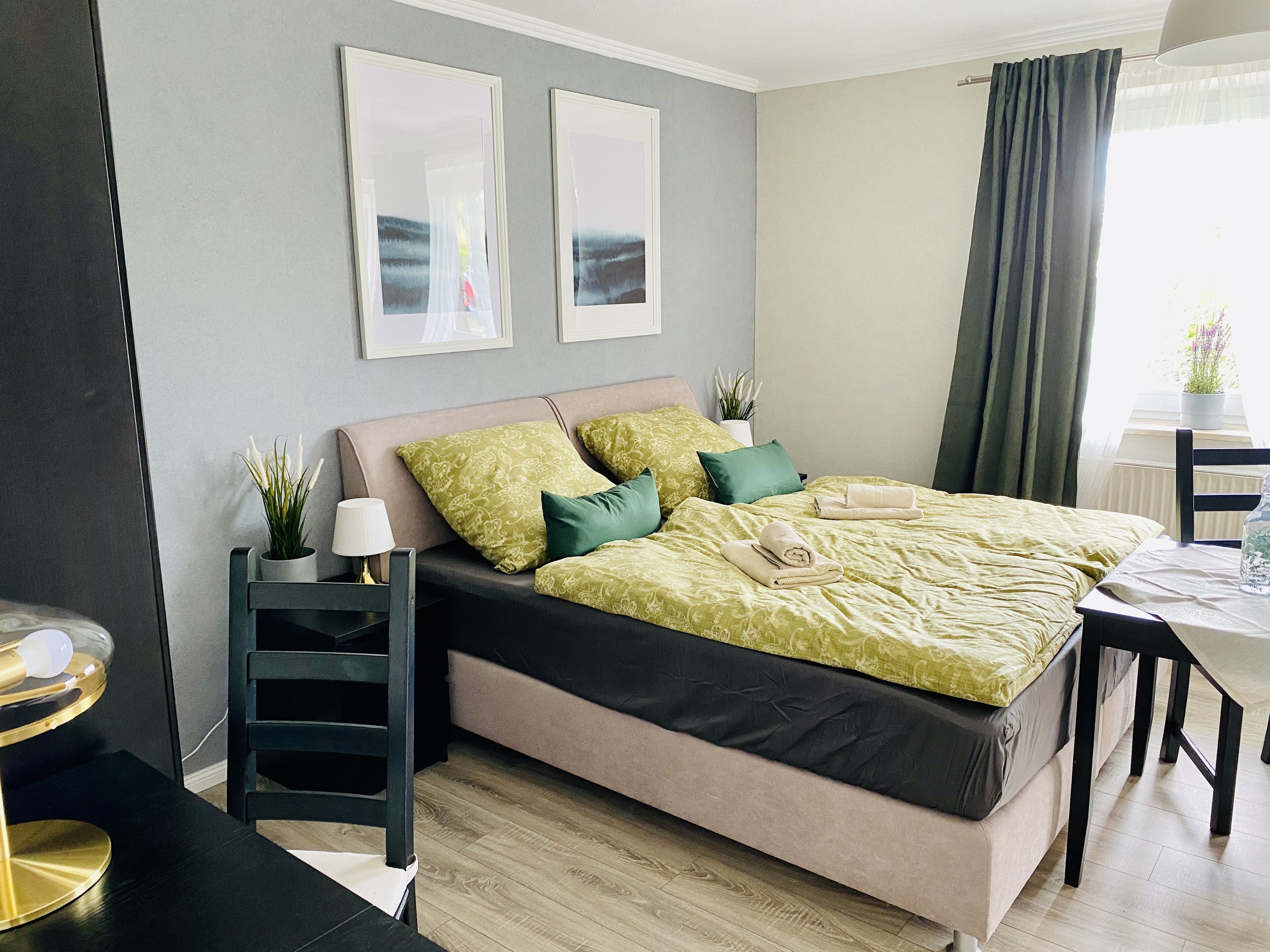 A neatly arranged bedroom with a bed, bedside table, lamp, and a view of the outdoors through the window.