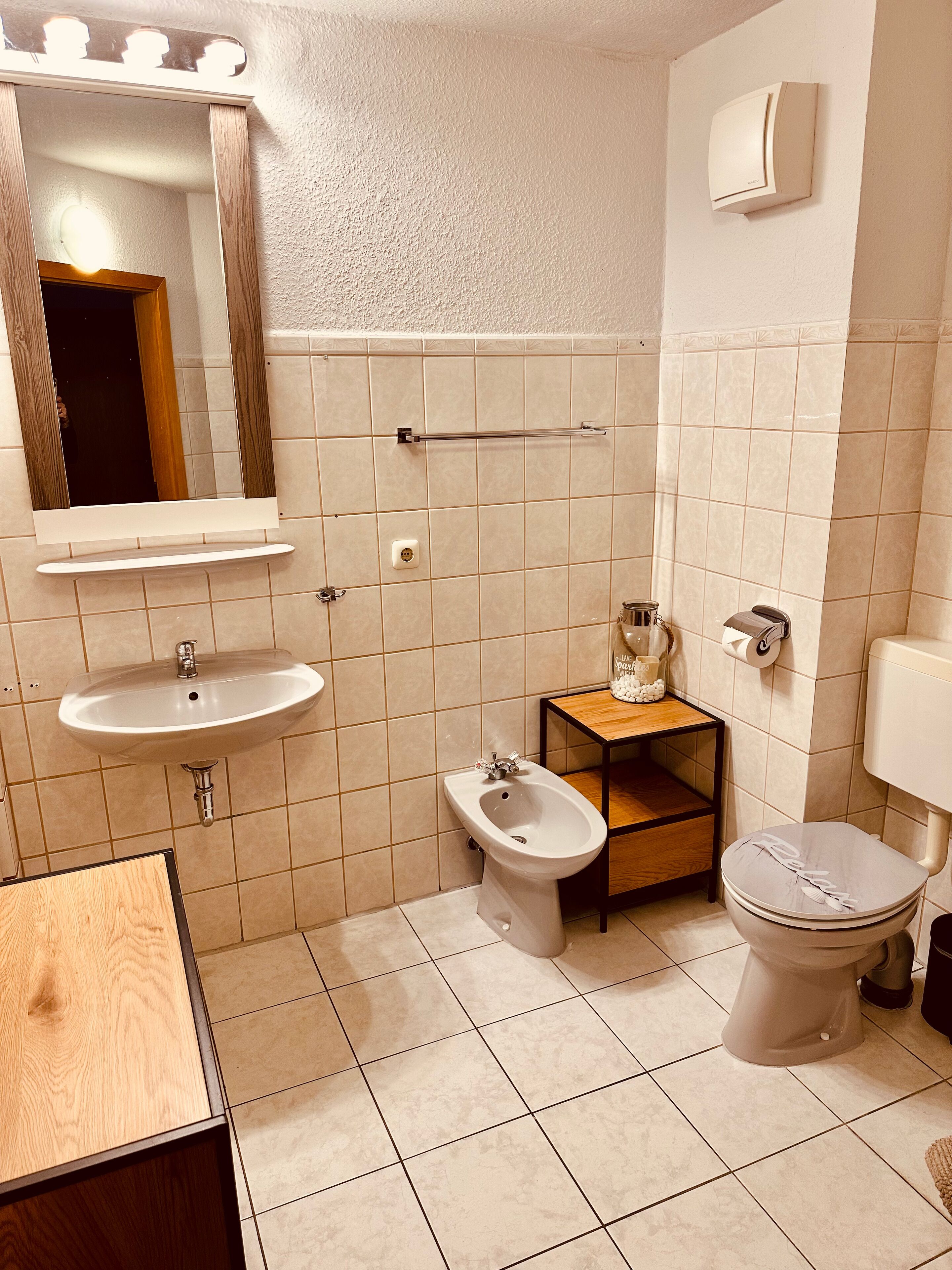 A bathroom with a sink, bidet, toilet, vanity, and a mirror with lights.