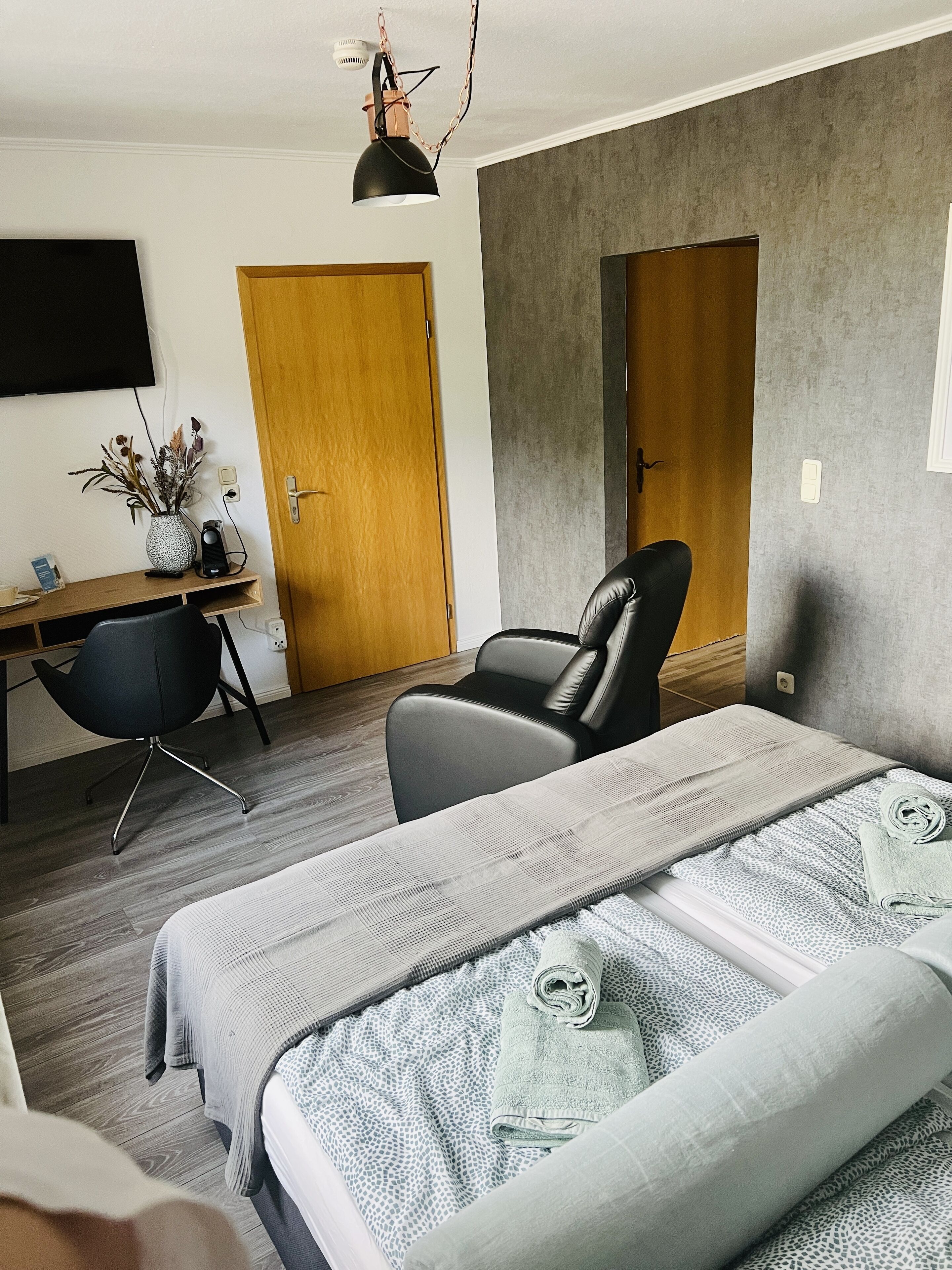 Una habitación de hotel moderna con una cama grande, un escritorio con silla, una mesita con un florero y dos puertas.