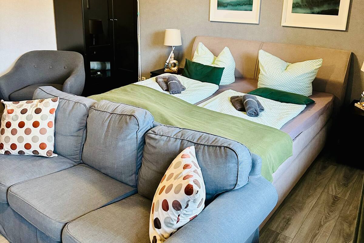 A modern hotel room with a large grey sofa, a bed with green and white bedding, and two framed landscape pictures on the wall.