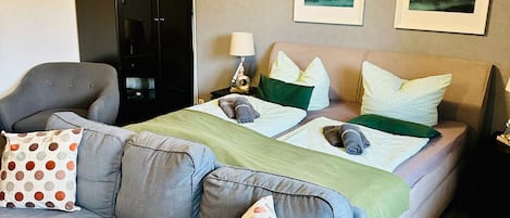 A modern hotel room with a large grey sofa, a bed with green and white bedding, and two framed landscape pictures on the wall.