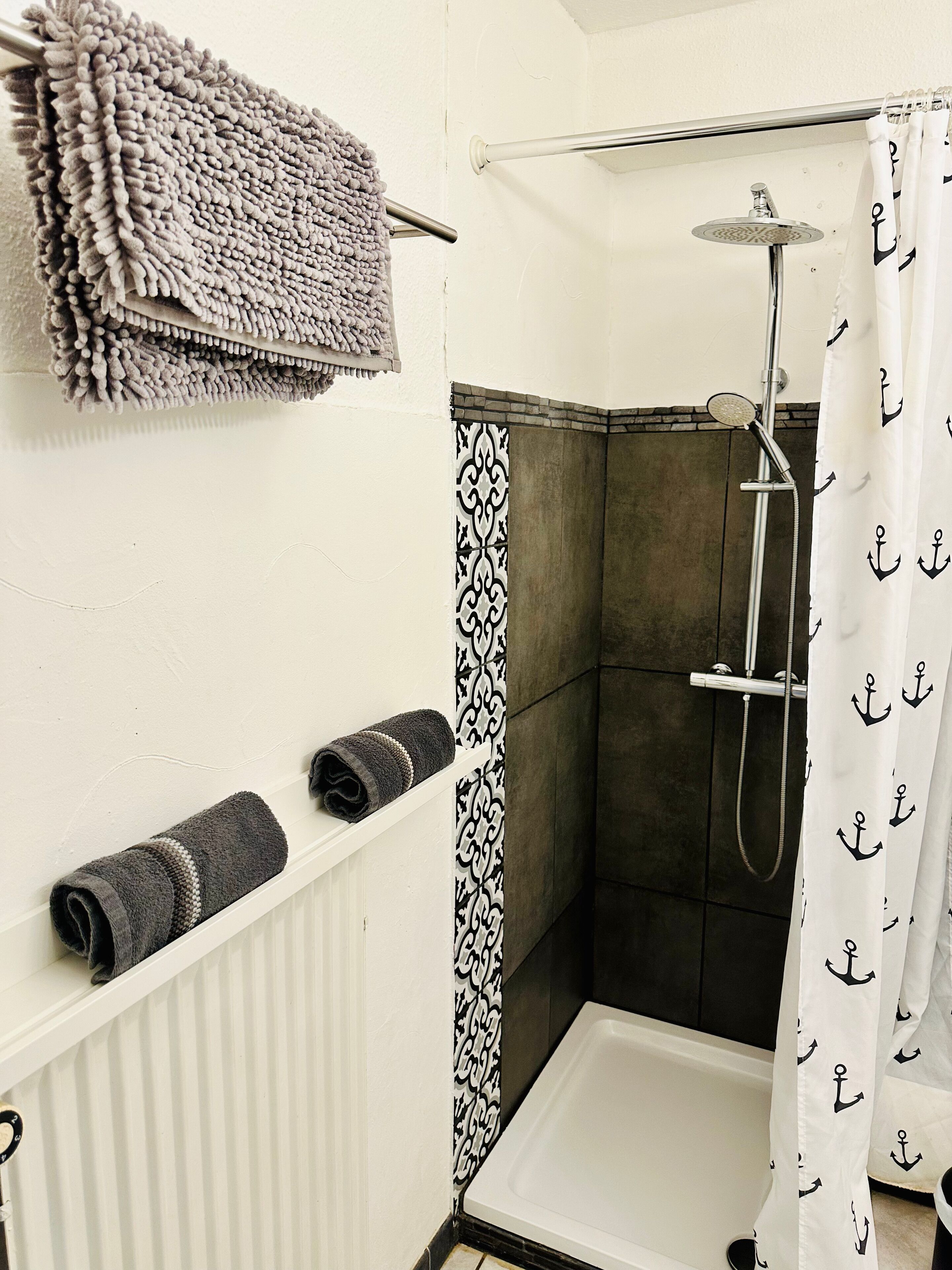 Zona de ducha con bañera blanca, cabezal de ducha y cortina con motivos de anclas. Hay toallas en un estante y un panel de pared estampado.