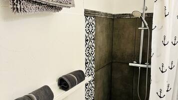 Un espace douche avec une baignoire blanche, une pomme de douche et un rideau orné de motifs d’ancres. Des serviettes sont posées sur une étagère, et un panneau mural décoratif orne le mur.