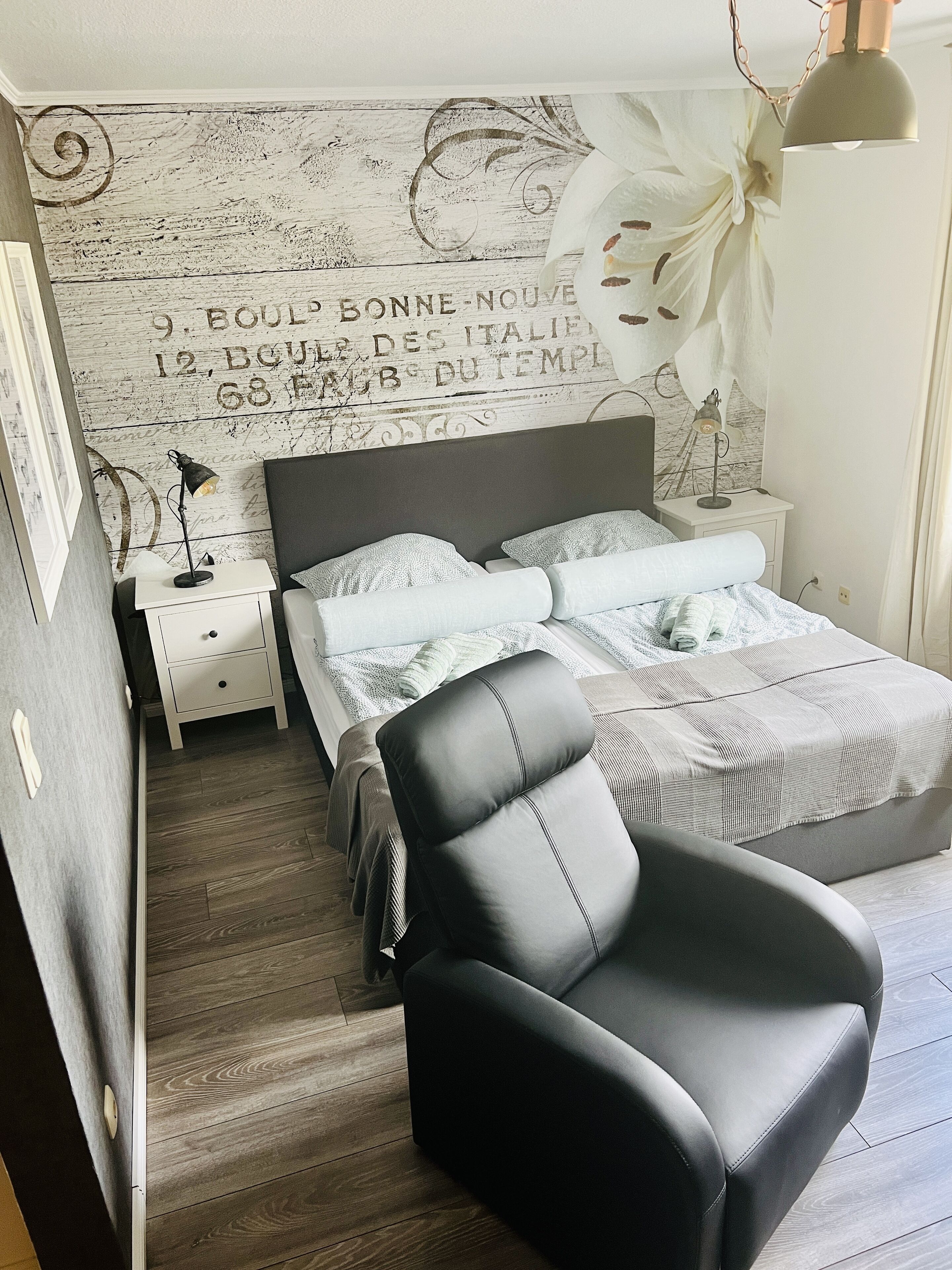 Une chambre à coucher comprenant un lit, une table de chevet, une lampe, une chaise et un mur orné d’un motif décoratif.