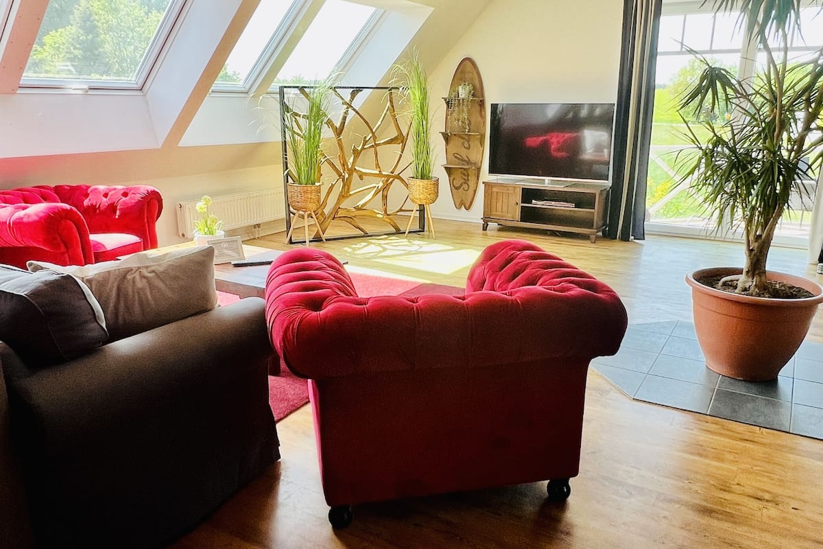 A modern living room with a red sofa, a brown sofa, and a wooden coffee table. There is a TV, a potted plant, and a decorative wall piece.