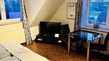 A room with a bed, a desk, a chair, and a TV. There are windows with curtains, a radiator, and a picture on the wall.