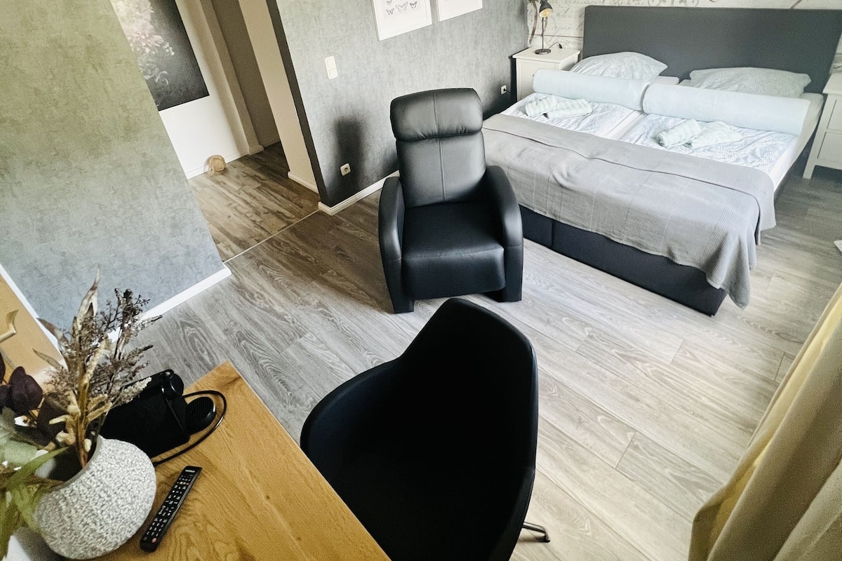 A modern hotel room with a bed, a desk, a chair, and a small table with a vase and remote control.