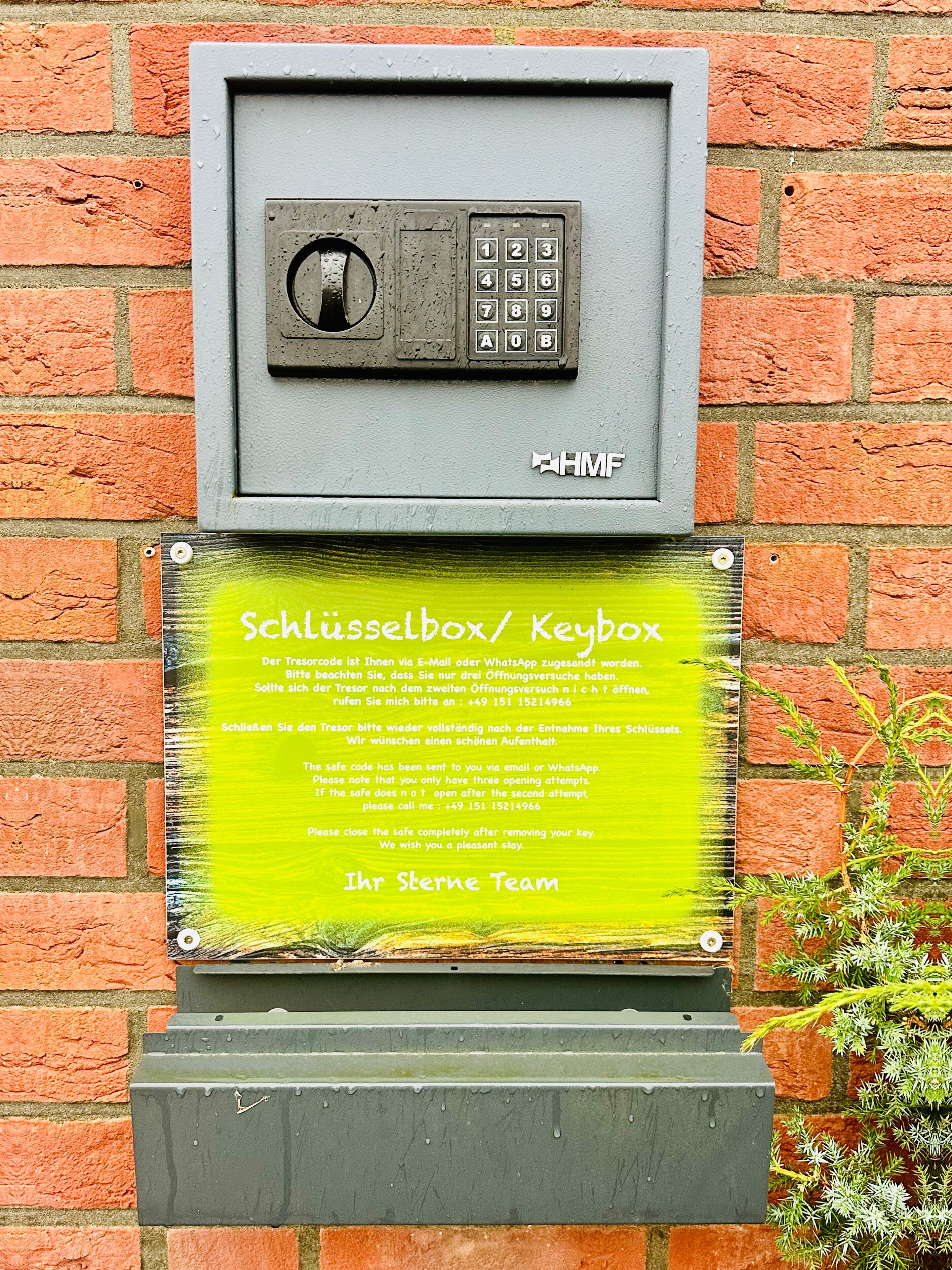 A keypad lockbox mounted on a brick wall with a green plaque below it.