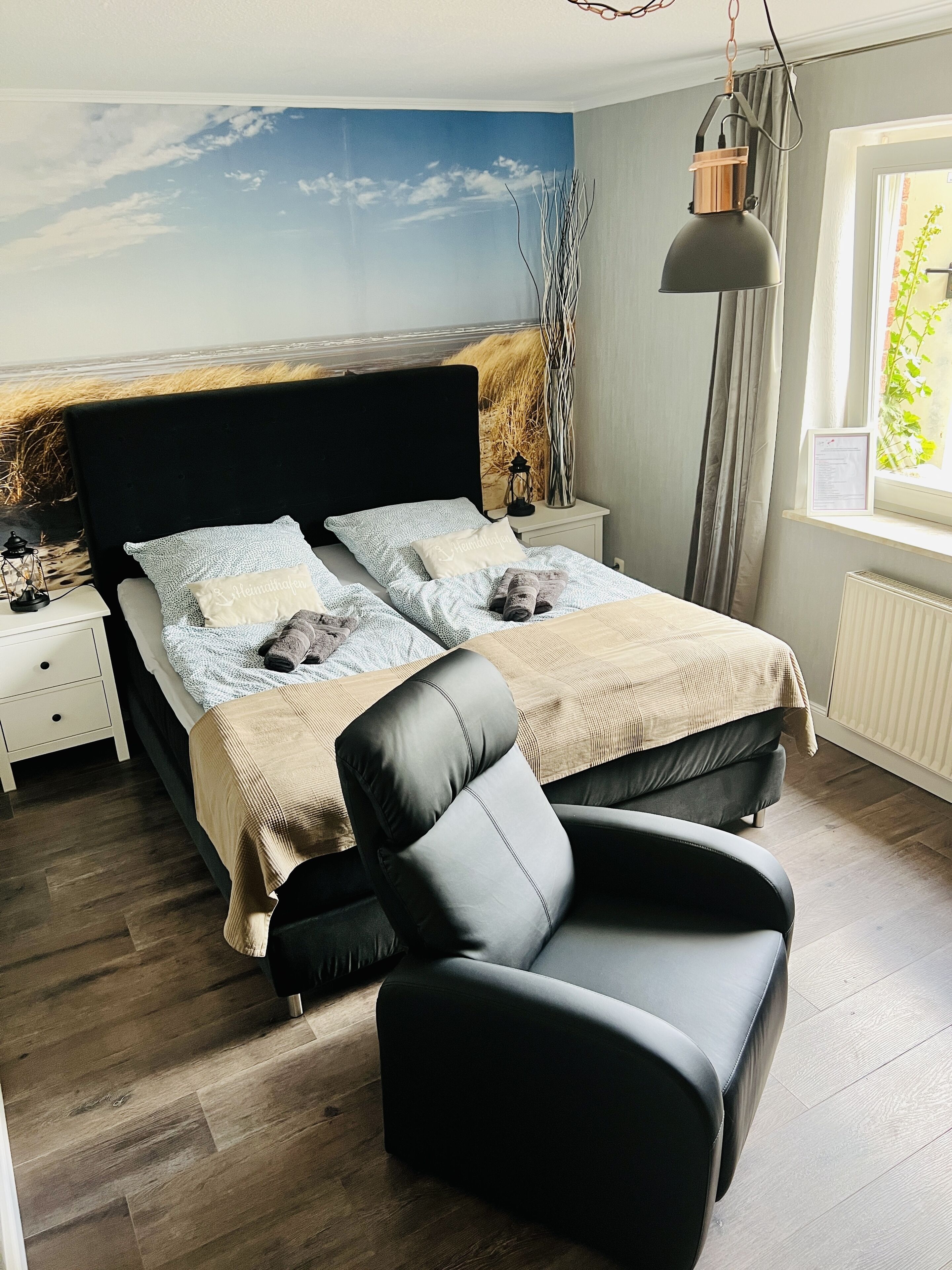 Een slaapkamer met een groot bed, twee nachtkastjes, een zwarte leren fauteuil, een hanglamp en een raam met uitzicht op groen.
