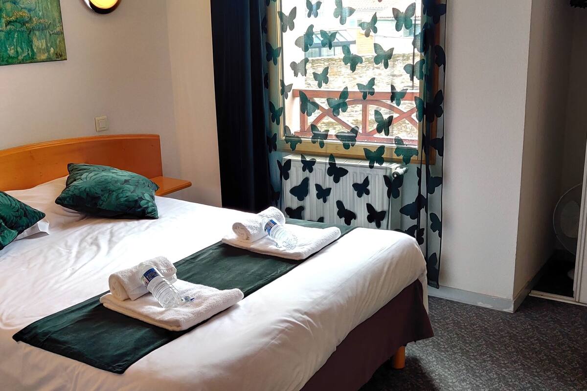 A hotel room with a bed, green pillows, a window with butterfly curtains, and a painting on the wall.