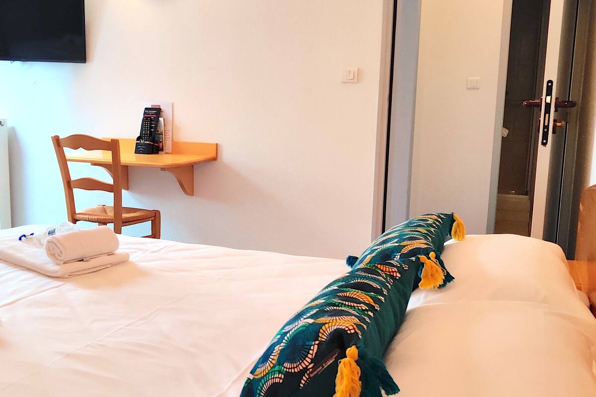 A hotel room with a bed, a chair, a small table with a remote control, and a window with curtains.