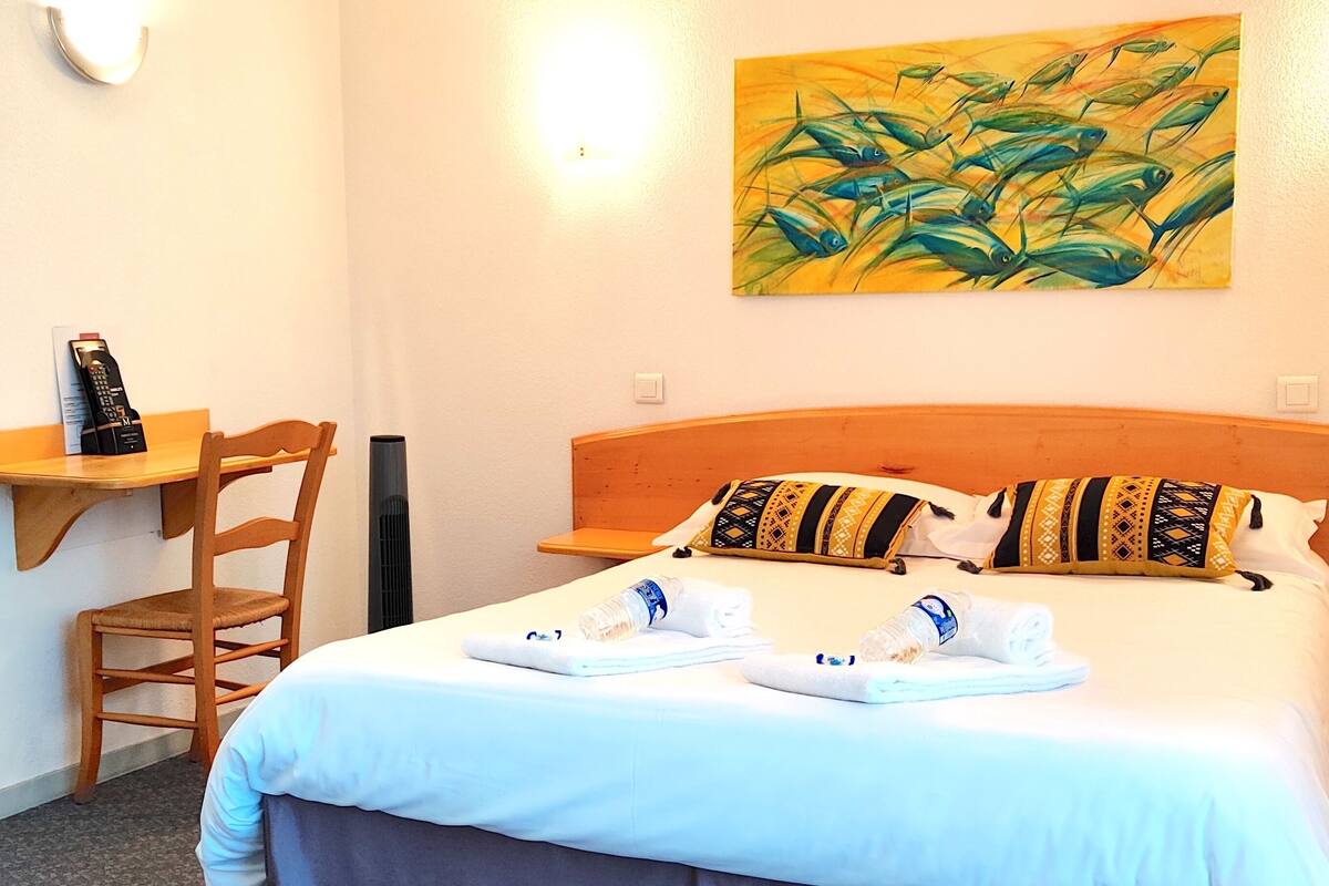 A hotel room with a bed, a chair, a small table, and a painting on the wall.