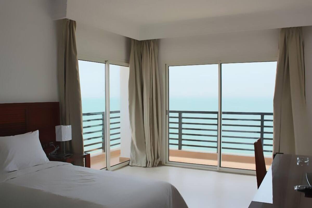 Una habitación de hotel con cama, escritorio y vistas al mar a través de amplios ventanales.