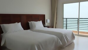 Habitación de hotel con dos camas, cabecera de madera, mesita de noche con lámpara y balcón con barandilla.