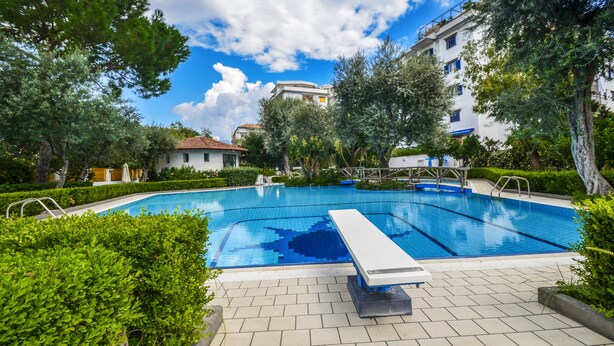 Piscina com trampolim, cercada por vegetação e edifícios.