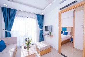 A modern hotel room with a sofa, a small table with a vase of flowers, a bed with blue pillows, and a TV on a wooden shelf. - Sincero Hotel & Apartment (Da Nang)