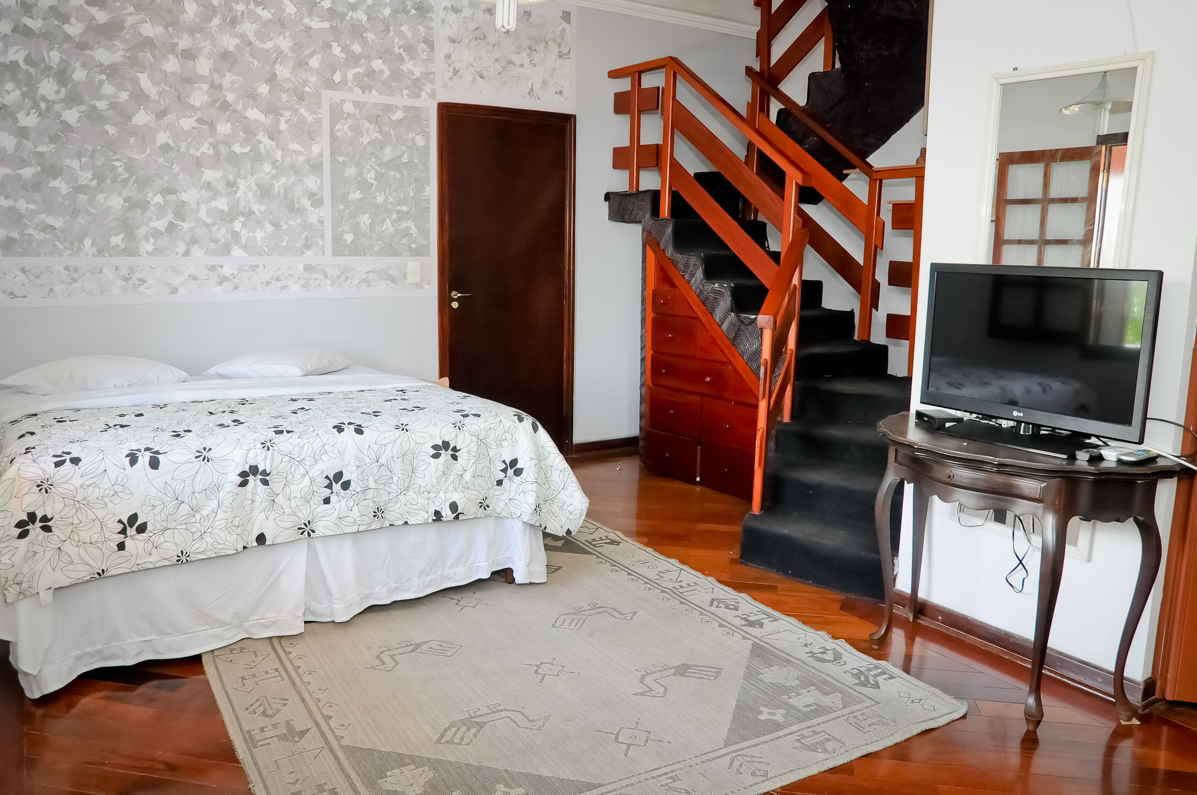 A bedroom with a bed, a TV on a stand, a wooden staircase, and a door.