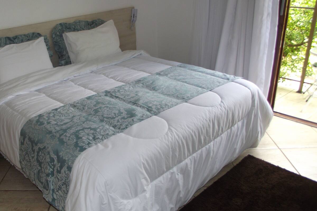 A neatly made bed with a white and blue patterned bedspread, a wooden headboard, and a view of greenery through a window.