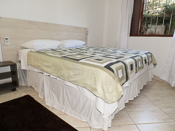 Una cama con una colcha estampada y almohadas blancas, una mesita de noche con un teléfono, y una ventana con vistas al follaje.