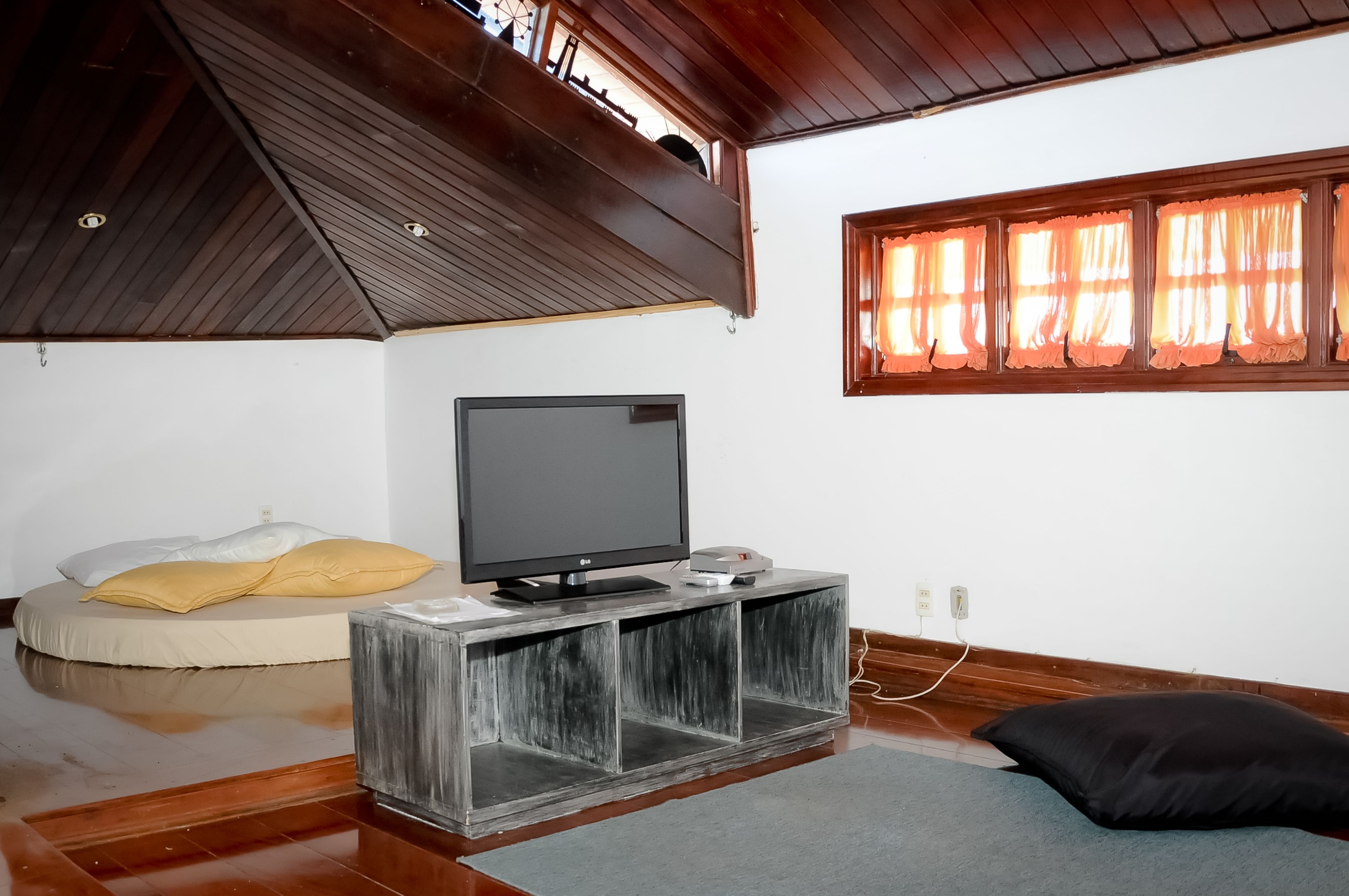 Une pièce avec un téléviseur posé sur un meuble en bois, un lit avec des oreillers, une fenêtre avec des rideaux et un plafond en bois.