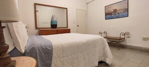 Un dormitorio con cama, cómoda, espejo, silla e imagen enmarcada en la pared.