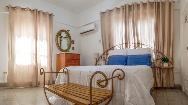 Un dormitorio con cama, banca de madera, cómoda, espejo y cortinas.