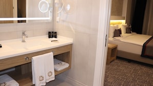 Un baño de hotel con un espejo grande, un lavabo y tocador. Hay un perchero con toallas y un estante pequeño con artículos de aseo.