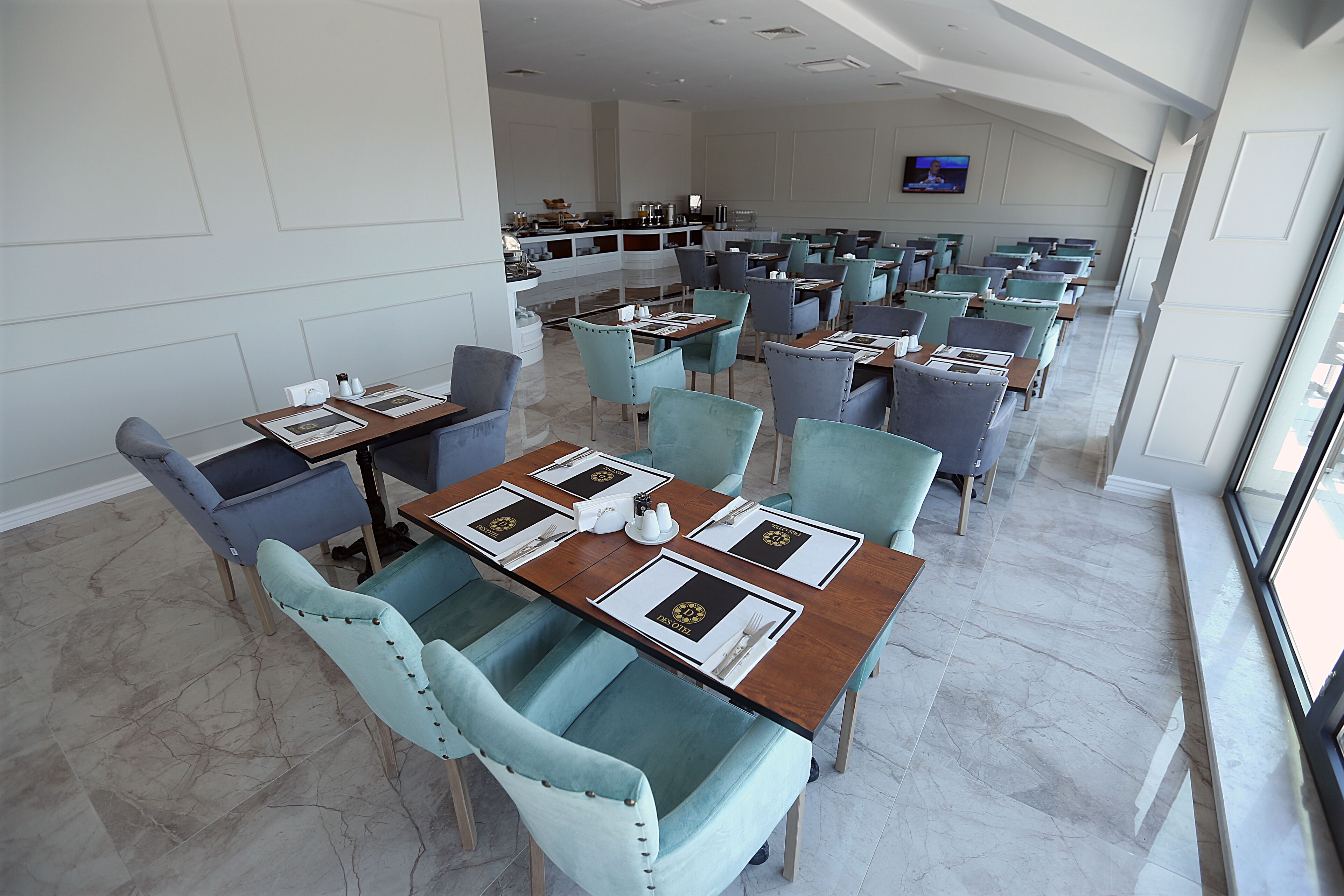 Un restaurante moderno con suelo de mármol, sillas azul verdosas y grises, y mesas ordenadas con individuales y cubertería.