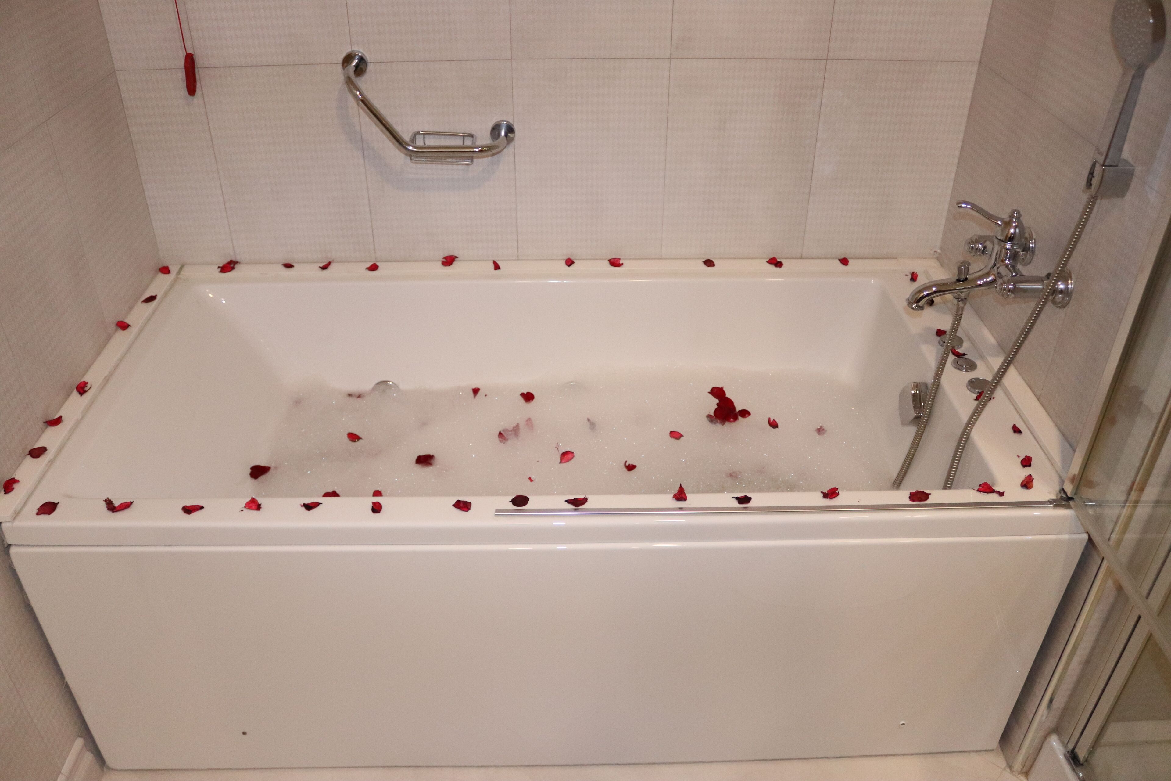 Una bañera blanca con pétalos de rosa esparcidos en su interior, un cabezal de ducha manual y un asidero en la pared.