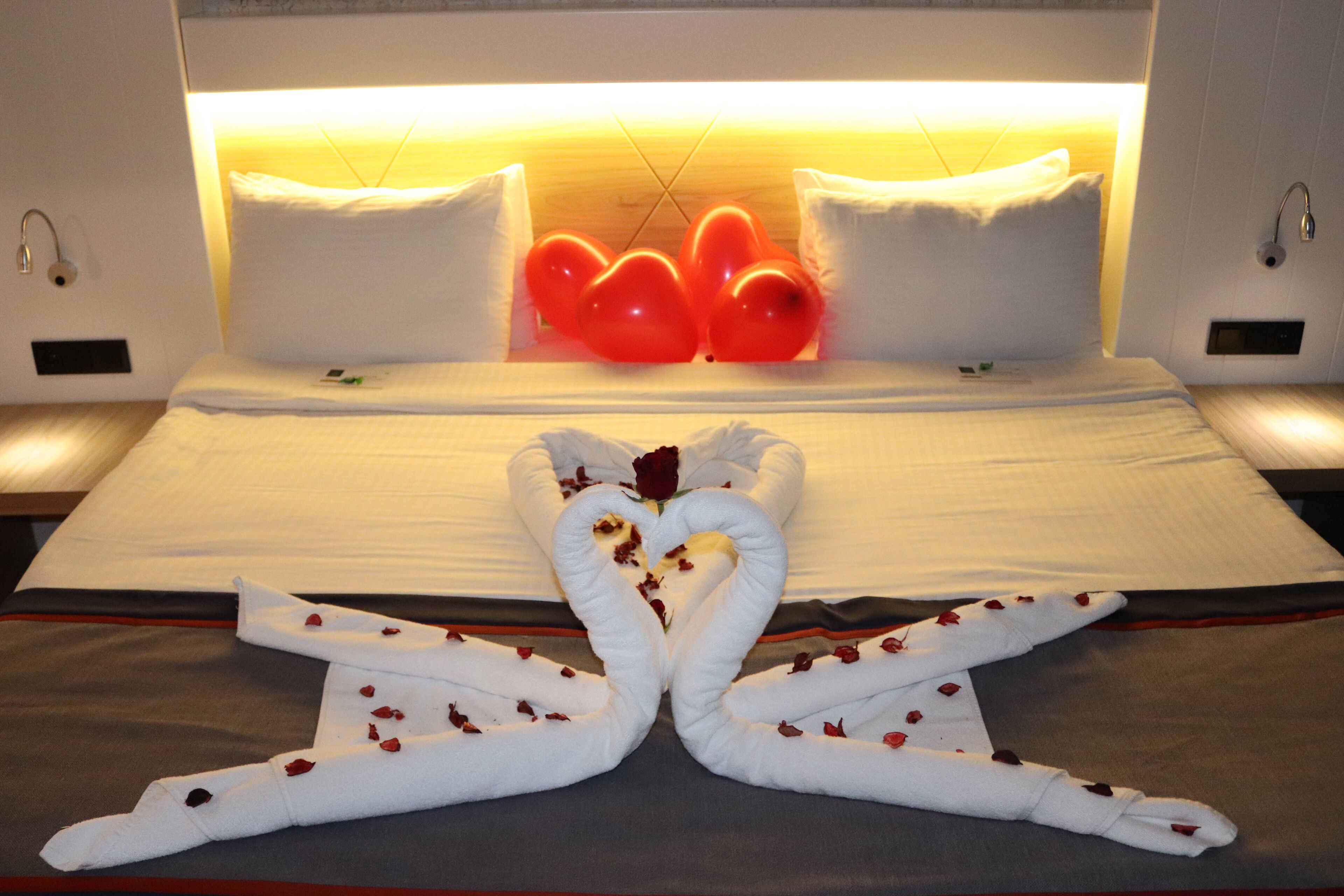 Habitación de hotel con cama, almohadas y una original disposición de toallas en forma de cisnes adornados con pétalos de rosa.