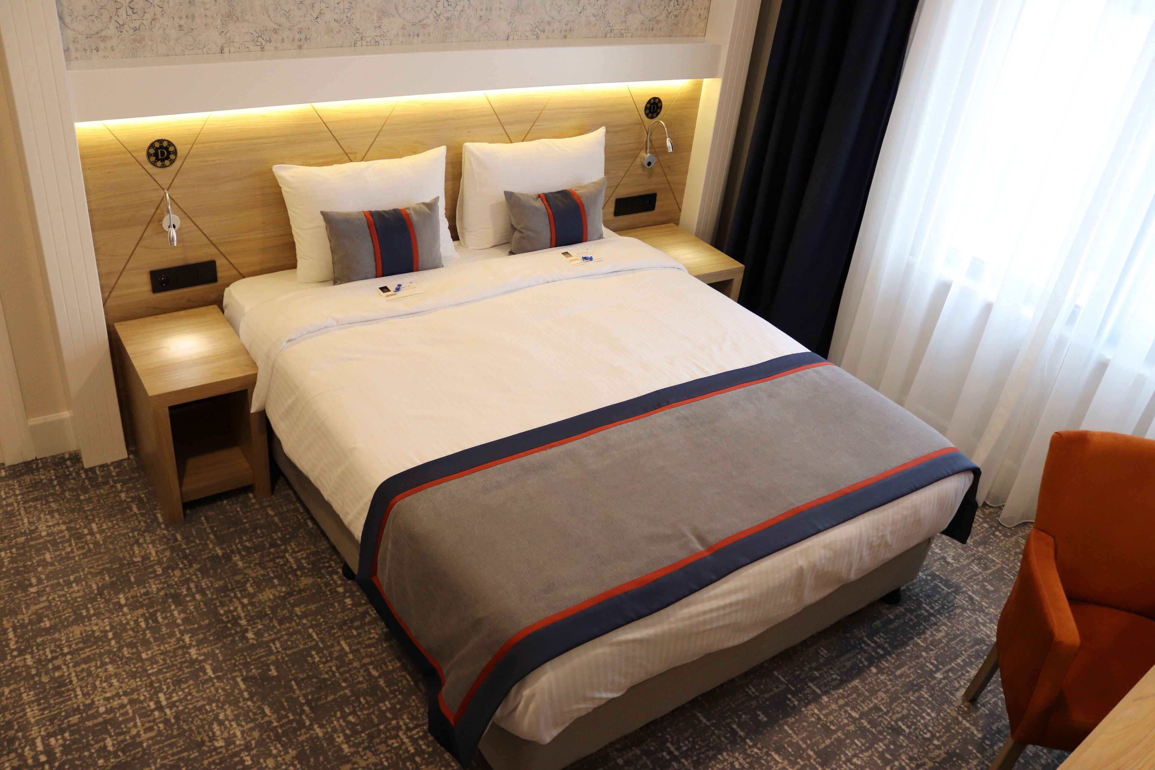 A hotel room with a large bed, two bedside tables, a nightstand, a chair, and a window with curtains.