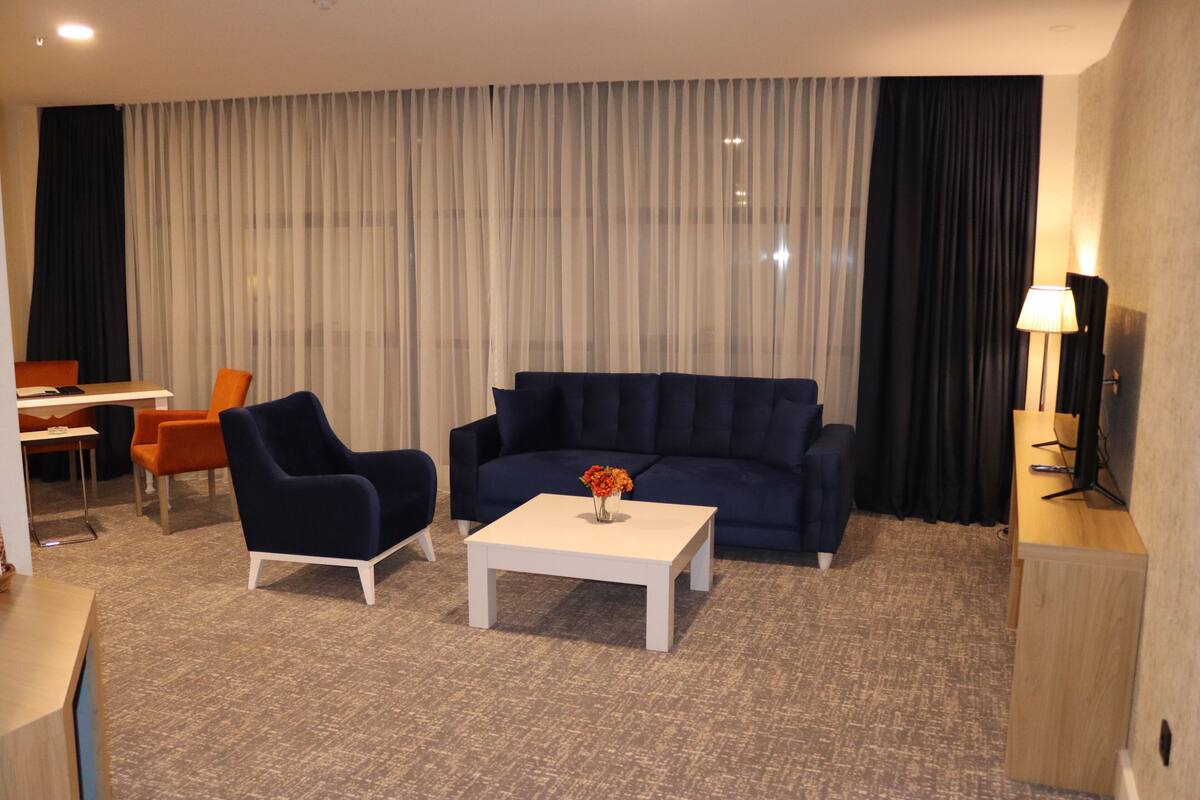 A hotel room with a dark blue sofa, a white coffee table with a vase of flowers, an orange armchair, a wooden desk, and a lamp.