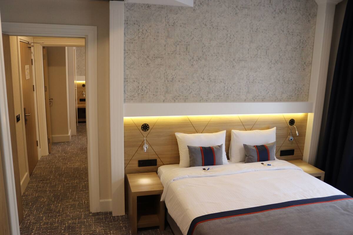 A hotel room with a large bed, a wooden headboard, a nightstand, and a wall-mounted light fixture.
