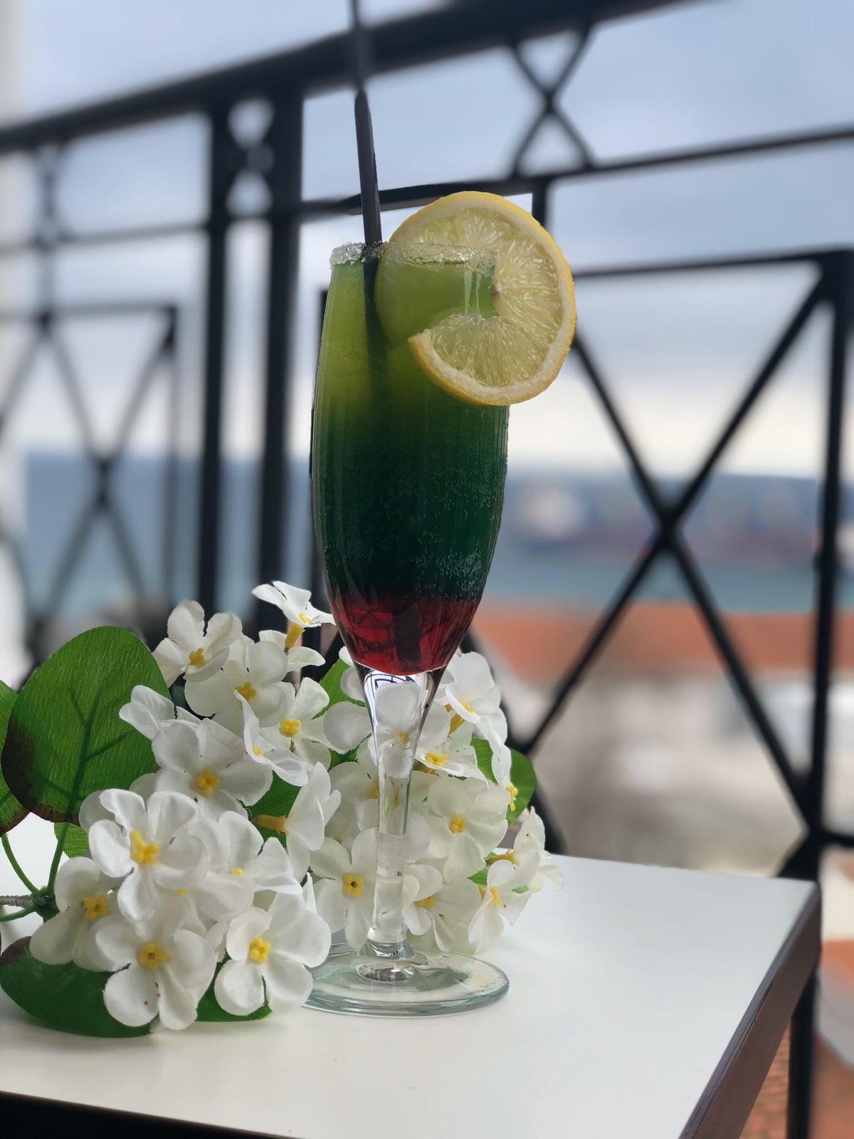 Un vaso de cóctel con líquido verde, una rodaja de limón y una pajita, sobre una mesa con flores blancas y una barandilla metálica al fondo.