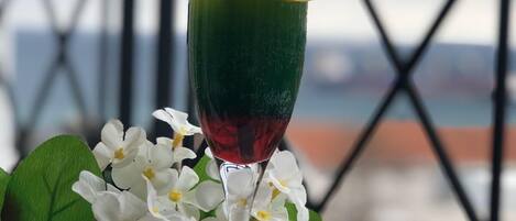 Un vaso de cóctel con líquido verde, una rodaja de limón y una pajita, sobre una mesa con flores blancas y una barandilla metálica al fondo.