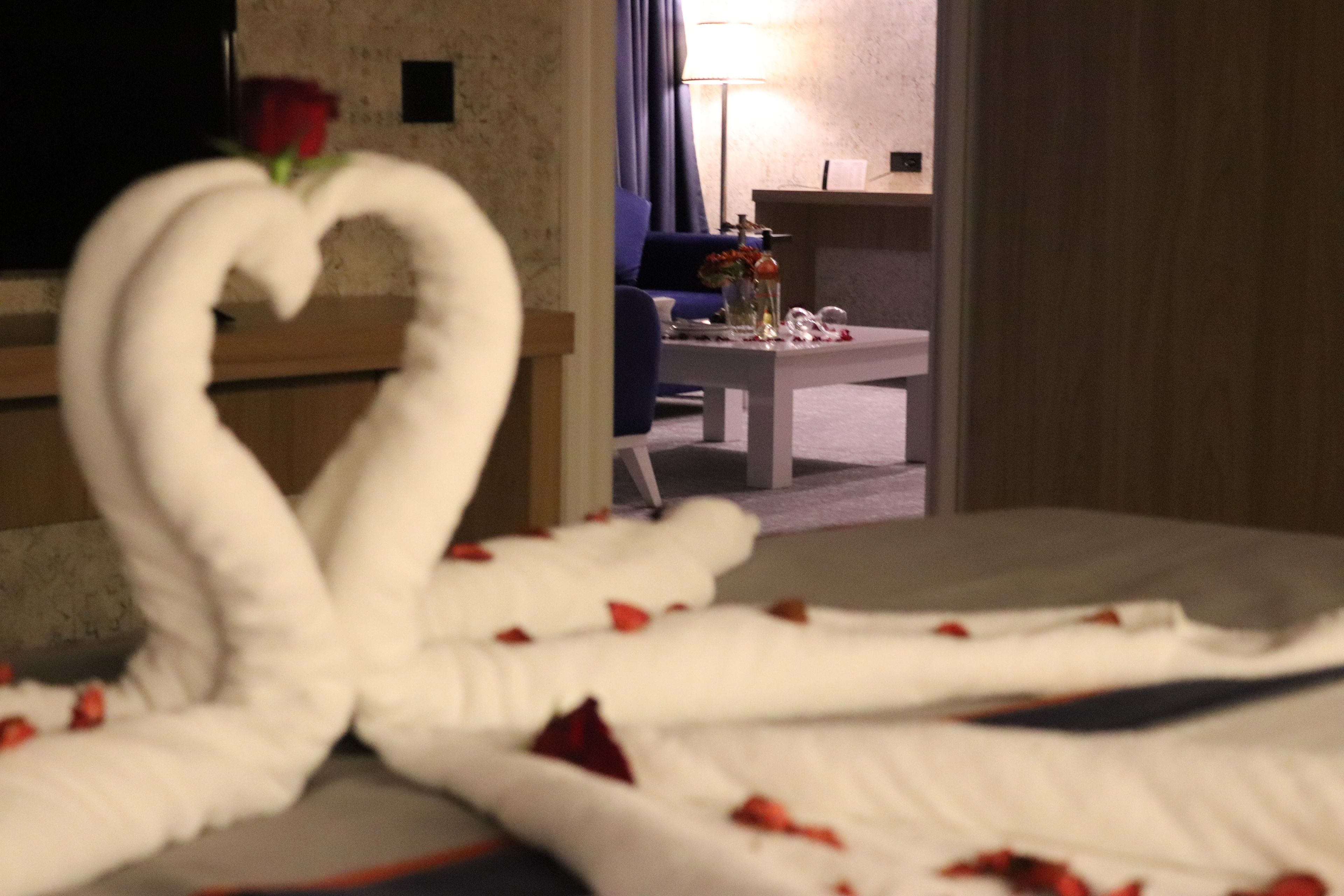 A bed with a heart-shaped towel arrangement and rose petals, a small table with a vase of flowers, and a blue sofa in a hotel room.