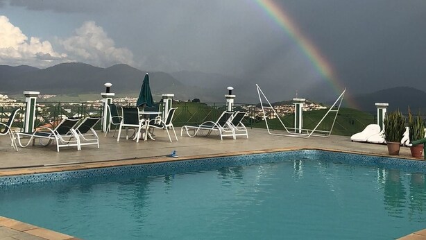 Una piscina con sillones de descanso y sombrillas, un arcoíris en el cielo, y una vista de montañas lejanas.