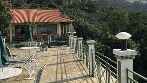 Una terraza al aire libre con mesas y sillas, un edificio con techo rojo y montañas al fondo.