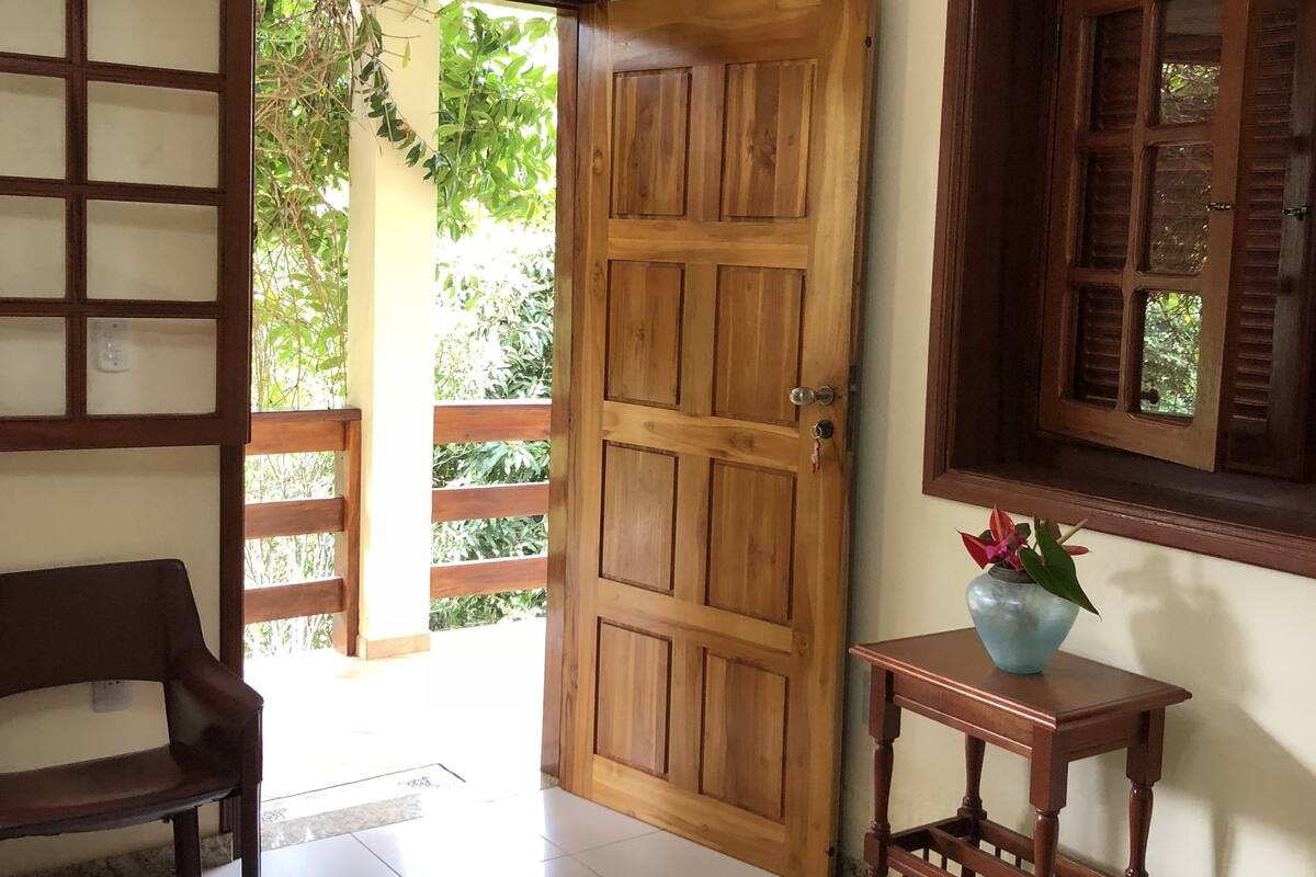 A room with a wooden door, a bench, a small table with a vase, and a window with shutters.